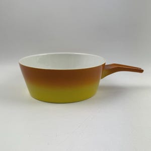 Puede incluir: Una cacerola vintage de dos tonos con asa. El interior es blanco, mientras que el exterior presenta un degradado de colores, pasando del amarillo al naranja. El asa es de color marrón. Ideal para cocinar.