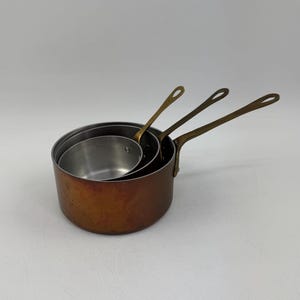 Op de afbeelding: Set van drie koperkleurige steelpannen met messing handvatten. De pannen zijn in elkaar genest, met verschillende maten. De binnenkant van de pannen is zilverkleurig, wat contrasteert met het warme, verouderde koper van de buitenkant.