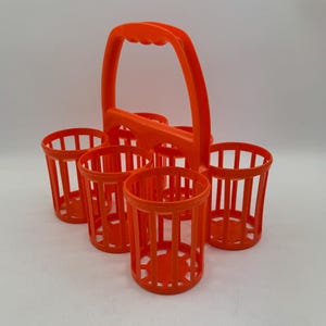May include: An orange plastic drinks carrier with a handle, designed to hold six individual cups. The carrier has a grid-like structure, with vertical bars forming the sides of each cup holder. The handle is arched for easy carrying.
