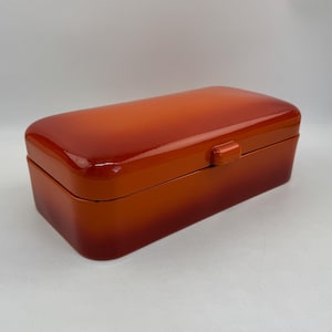 May include: A rectangular, glossy orange box with a rounded lid and a small clasp. The color gradient transitions from a lighter orange at the top to a darker shade at the base. The box is set against a plain white background.