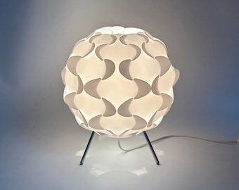 May include: A white table lamp with a unique, geometric, spherical shade made of overlapping, curved panels. The lamp has a silver metal base with three legs.