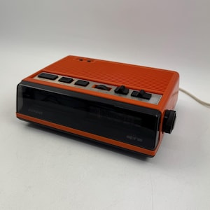 Könnte beinhalten: Ein orangefarbener und schwarzer Vintage-Wecker mit Digitalanzeige. Die Uhr hat mehrere Knöpfe und einen Drehregler an der Oberseite und an der Seite. Der Markenname "alpha" ist auf der Vorderseite sichtbar.