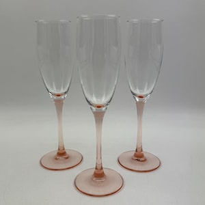 Op de afbeelding: Drie heldere champagneglazen met roze stelen en voetjes. De glazen zijn in een driehoekige formatie geplaatst tegen een effen witte achtergrond. De stelen en voetjes hebben een zachte, roze tint, wat een vleugje kleur toevoegt aan het elegante ontwerp.