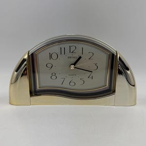 Op de afbeelding: Een goudkleurige Seiko quartz klok met een gebogen, doorzichtige plastic bovenkant. De wijzerplaat is wit met zwarte cijfers en wijzers. De klok is rechthoekig met afgeronde hoeken en een gouden basis.