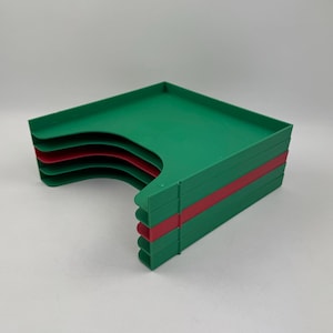 Puede incluir: Una pila de bandejas de plástico verdes y rojas. Las bandejas son rectangulares con un recorte curvo en un lado. Una franja roja acentúa el lado de la pila. Las bandejas están apiladas sobre una superficie blanca.