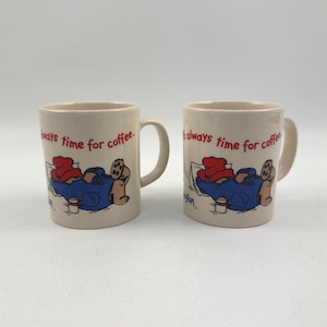 Op de afbeelding: Twee beige koffiemokken met een cartoonillustratie van een beer met een rode hoed en blauwe jas, met de tekst "always time for coffee". De beer ligt en houdt een koffiekopje vast. De mokken hebben een wit handvat.