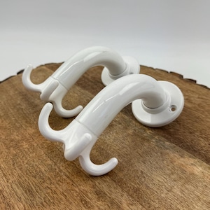 Op de afbeelding: Twee witte, gebogen kapstokken met een drievoudig haakontwerp. De haken zijn gemonteerd op een cirkelvormige basis met schroefgaten. De haken worden gepresenteerd op een houten oppervlak.