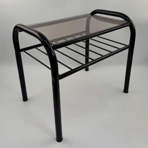 Puede incluir: Una pequeña mesa auxiliar de metal negro con un marco rectangular, patas curvas y una superficie superior de cristal tintado en marrón. Debajo de la parte superior de cristal, la mesa incluye un estante de listones metálicos.