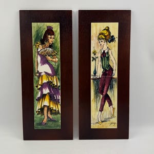 May include: Two rectangular wall plaques featuring vertical ceramic tiles mounted on dark wood. The left tile depicts a person in a purple and yellow ruffled dress holding a fan. The right tile shows a person in a green vest and maroon trousers.