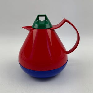 Op de afbeelding: Een retro-stijl, rood en blauw geïsoleerde thermoskan met een groene deksel en een rode handgreep. De thermoskan heeft een afgeronde vorm en een schenktuit. Het ontwerp is eenvoudig en kleurrijk, doet denken aan vintage keukengerei.
