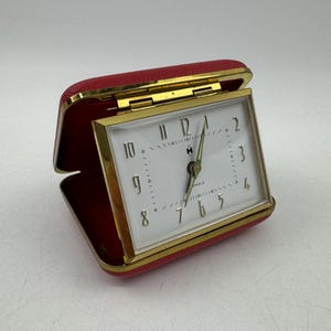 Könnte beinhalten: Ein Vintage-Reisewecker in einem roten Kunstleder-Etui. Das Zifferblatt ist weiß mit goldfarbenen Zeigern und Ziffern. Das Etui ist geöffnet und zeigt das Zifferblatt und einen goldfarbenen Rahmen. Misst etwa 7.6 cm breit.