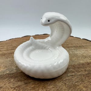 Op de afbeelding: Een witte keramische cobra-figuur met zwarte ogen, opgerold in een verdedigende houding. De slang staat op een houten oppervlak, waarbij de gladde, glanzende afwerking wordt getoond. Het ontwerp is eenvoudig maar opvallend.