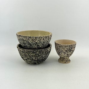 Puede incluir: Tres cuencos de cerámica con un esmalte moteado en blanco y negro. Dos cuencos están apilados uno encima del otro, y el tercer cuenco es más pequeño y tiene una base con pie.