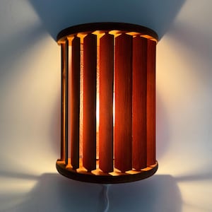 May include: A cylindrical wall lamp featuring vertical dark wood slats arranged around a central light source. The design includes a circular top and bottom frame, emitting a warm glow through the gaps between the slats, with a white power cord.