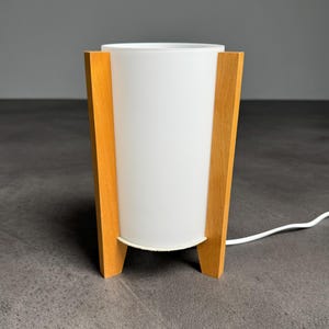 Op de afbeelding: Een cilindrische tafellamp met een witte matglazen kap die wordt vastgehouden door twee lichtgekleurde houten steunen. De lamp staat rechtop en is voorzien van een wit elektrisch snoer dat uit de voet komt, voor een strak en functioneel ontwerp.