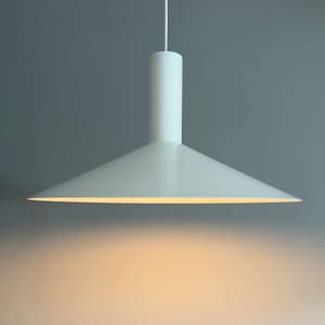 May include: A matte white pendant light fixture featuring a wide, conical shade and a cylindrical top mount. The lamp is suspended by a thin white cord and emits a warm, soft glow from underneath, illuminating the space directly below the shade.