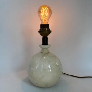 Op de afbeelding: Een vintage tafellamp met een bolvormige basis met een marmer effect. De basis is crèmewit met een wervelend patroon. De lamp heeft een decoratieve gloeilamp die brandt en een warm, oranje licht uitstraalt. Een zwarte kabel komt uit de basis.