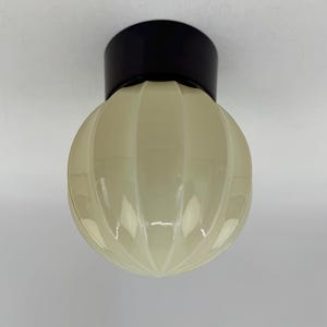Op de afbeelding: Een plafondlamp met een bolvormige, geribbelde glazen kap in een zachte crèmekleur. De kap is bevestigd aan een massieve zwarte ronde voet die direct aan het plafond wordt gemonteerd, wat zorgt voor een strak en eenvoudig profiel.