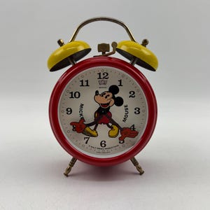 May include: A vintage red and yellow Mickey Mouse alarm clock. The clock face features Mickey Mouse and the words "Mickey Mouse". The clock has a metal frame and legs. The clock's bells are yellow.