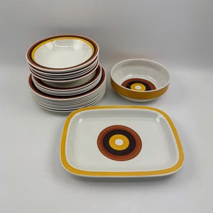 Op de afbeelding: Een verzameling vintage keramisch serviesgoed met een retro design. De set bevat kommen en borden met een witte basis en concentrische cirkels in bruin, geel en oranje. De borden zijn rechthoekig met afgeronde hoeken.