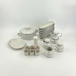 Può includere: Un set di stoviglie da cucina in ceramica bianca con finiture nere. Il set comprende una casseruola con coperchio, un piatto da portata, un saliera, sei piccole tazze, due piccole brocche e una pila di sei piatti da portata rettangolari.