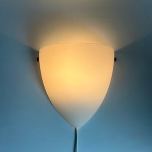 Op de afbeelding: Een wandlamp met een witte, schildvormige kap. Het licht straalt warm uit en de lamp is bevestigd aan een lichtblauwe muur. Een wit snoer loopt van de onderkant.