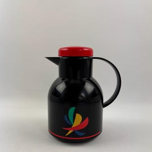 Op de afbeelding: Zwarte thermoskan met een rood deksel en een kleurrijk abstract ontwerp. Het ontwerp heeft een gestileerde werveling van groen, geel, rood en blauw.