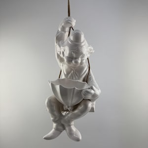Op de afbeelding: Witte keramische figuur van een clown die een paraplu vasthoudt, hangend aan een touw. De clown draagt ​​een hoed en heeft een grillige uitdrukking.