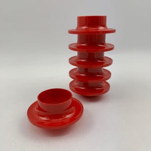 May include: Bright red, stackable, plastic container set. The set includes a base with a small, round bowl and a tall, segmented tower. The tower is composed of several disc-shaped layers, creating a unique, modern design. The pieces are arranged on a white surface.