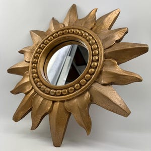 Op de afbeelding: Een decoratieve zonne-spiegel met een gouden afwerking. De spiegel heeft een ronde kern omgeven door een kralenrand en zonnestraalvormige randen. Ontworpen om aan de muur te worden bevestigd.