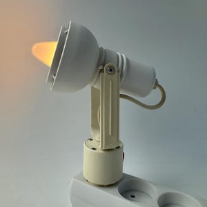 May include: A cream-coloured spotlight lamp emitting a warm yellow glow. The lamp is connected to a white power strip with two sockets. The design is simple and functional, with an adjustable head.