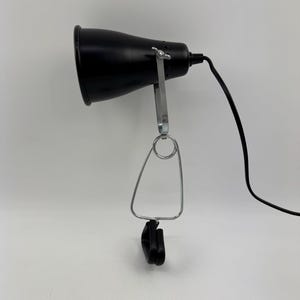 Könnte beinhalten: Eine schwarze Schreibtischlampe mit konischem Schirm. Die Lampe hat einen silberfarbenen, verstellbaren Arm und eine Klemme zur Befestigung. Ein schwarzes Netzkabel geht von der Lampe ab.