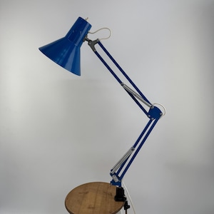 Könnte beinhalten: Eine blaue Schreibtischlampe mit konischem Schirm und verstellbarem Arm ist an einer Holzoberfläche befestigt. Die Lampe wird von einem weißen Hocker getragen. Der Arm der Lampe erstreckt sich auf etwa 61 cm.