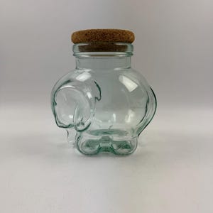 May include: A clear glass jar shaped like an elephant with a cork lid. The jar is empty.