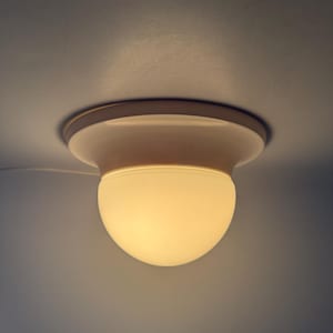 May include: A flush-mount ceiling light fitting with a rounded, opaque glass shade. The fitting is off-white and emits a warm glow. The light is mounted on a white ceiling.