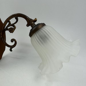 May include: An antique wall sconce with a frosted glass shade shaped like a flower. The sconce has a decorative bronze-colored metal arm and a wooden backing. The light fixture is designed to be mounted on a wall.