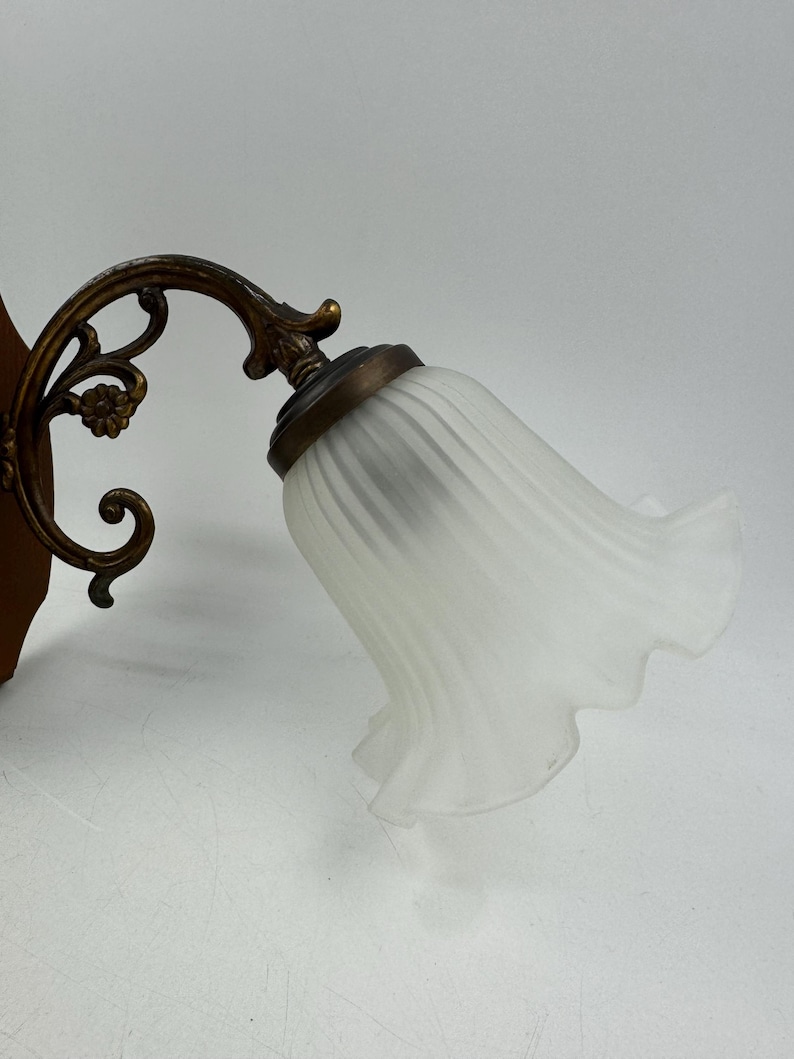 May include: An antique wall sconce with a frosted glass shade shaped like a flower. The sconce has a decorative bronze-colored metal arm and a wooden backing. The light fixture is designed to be mounted on a wall.