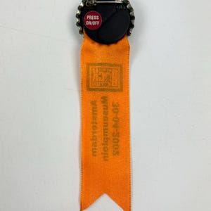 Puede incluir: Cinta naranja con un pin de tap&oacute;n de botella de metal. El tap&oacute;n de la botella tiene un bot&oacute;n rojo que dice "PRESS ON/OFF". La cinta tiene el texto "2003-2007 Mid-museum, metropolitan" impreso en ella.