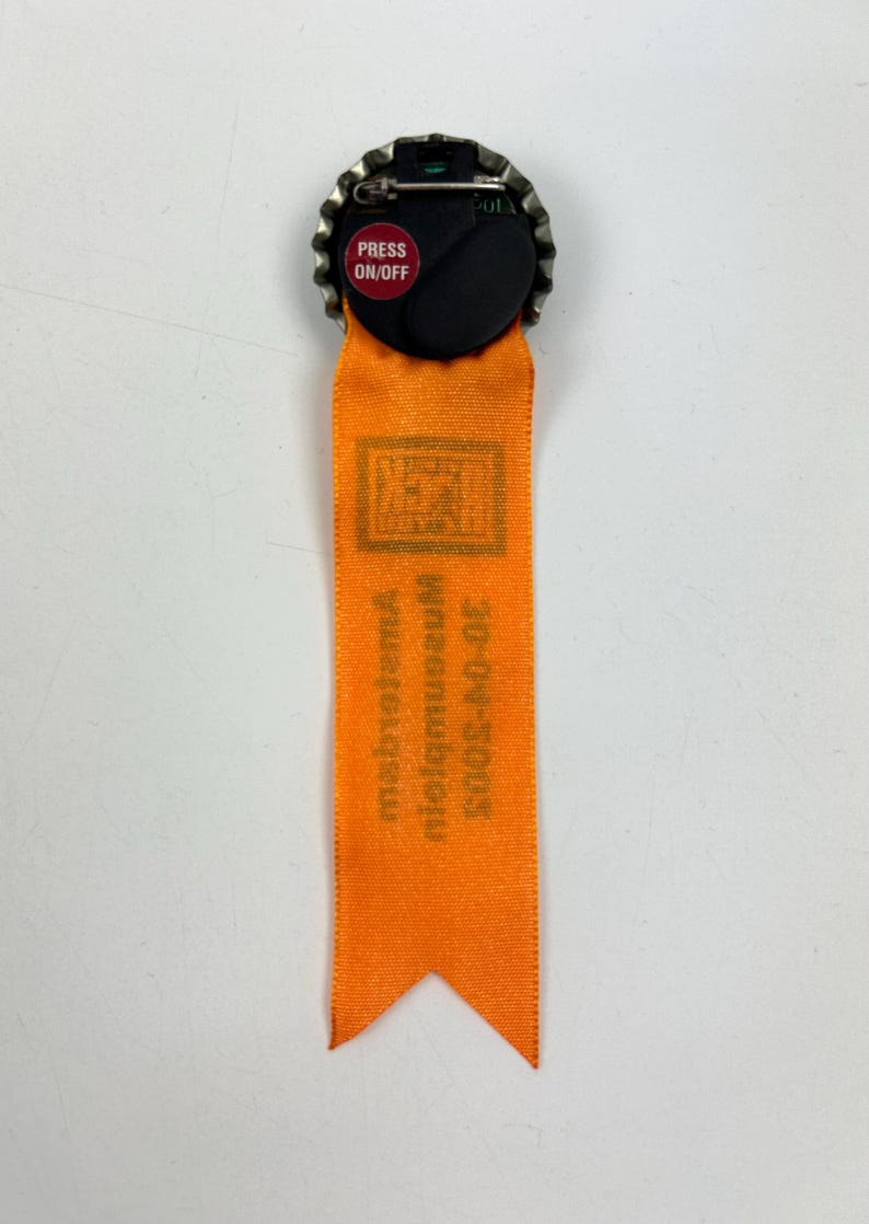 Puede incluir: Cinta naranja con un pin de tap&oacute;n de botella de metal. El tap&oacute;n de la botella tiene un bot&oacute;n rojo que dice "PRESS ON/OFF". La cinta tiene el texto "2003-2007 Mid-museum, metropolitan" impreso en ella.