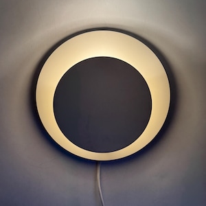 May include: A contemporary circular wall light emitting a warm glow. The light features a white outer ring and a dark grey centre. A white power cable extends from the base. The light is mounted on a grey wall.