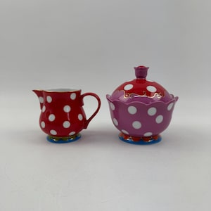 Puede incluir: Un juego de crema y azucarero de lunares rojos y blancos. La crema tiene un asa y un pico. El azucarero tiene una tapa con un asa en forma de rosa. Ambas piezas tienen un borde azul y dorado.