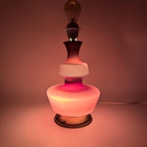 May include: A pink and white glass table lamp with a gold base. The lamp has a round, layered design and is lit with a light bulb.
