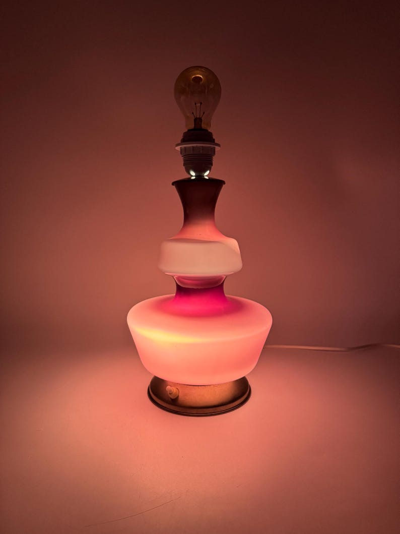 May include: A pink and white glass table lamp with a gold base. The lamp has a round, layered design and is lit with a light bulb.
