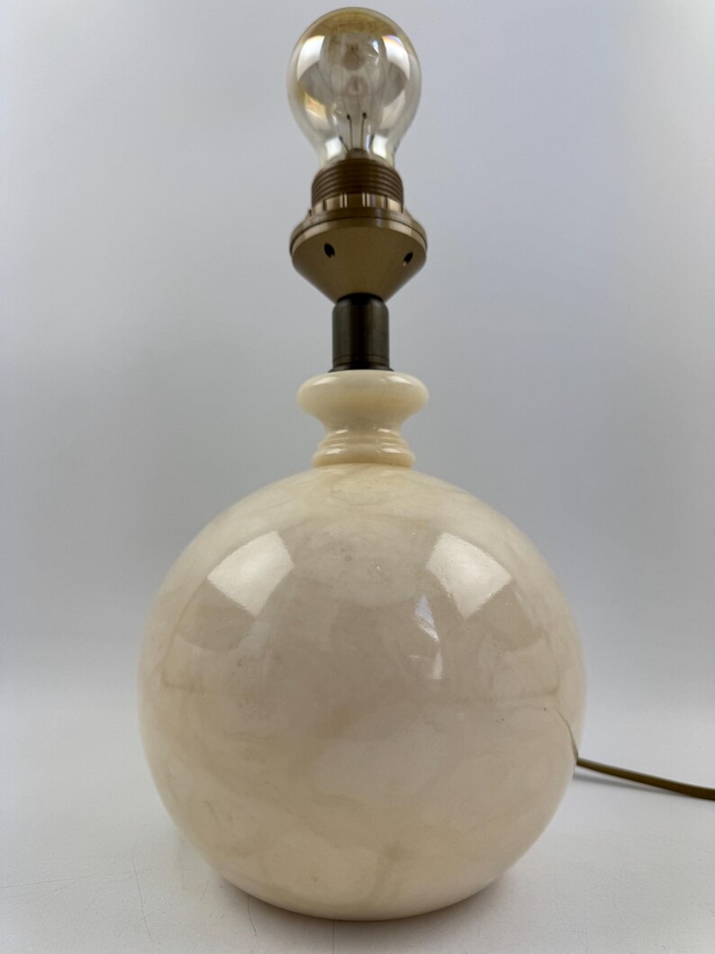 Peut inclure: Une lampe de table sph&eacute;rique cr&egrave;me avec une ampoule de style vintage. La lampe a une base ronde et une douille de couleur laiton. L'ampoule est transparente avec un filament visible. Un cordon &eacute;lectrique marron s'&eacute;tend de la base.