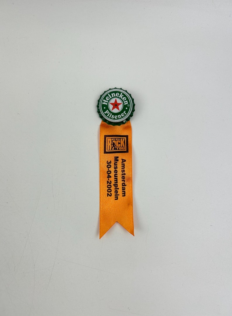 Puede incluir: Un tap&oacute;n de botella Heineken Pilsener verde y blanco con una estrella roja unida a una cinta naranja con el texto "We Will Rock You" y "Amsterdam Museumplein 30-04-2002".