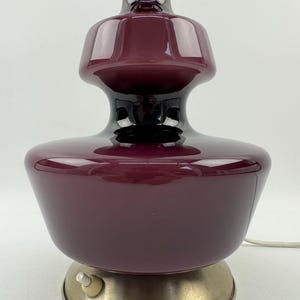 May include: A burgundy glass table lamp with a metal base and a cord with an inline switch. The lamp has a unique, layered design.