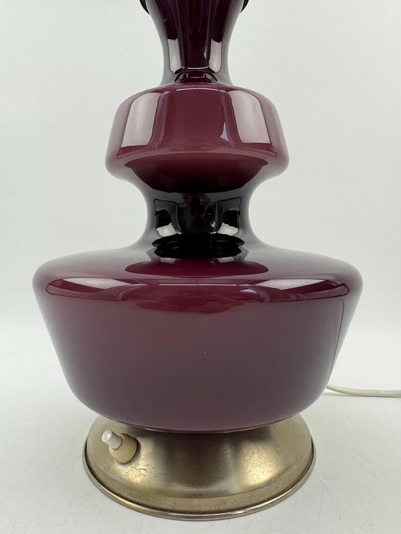 May include: A burgundy glass table lamp with a metal base and a cord with an inline switch. The lamp has a unique, layered design.