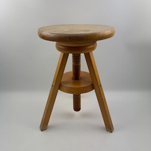 Pode incluir: Um banco ajustável de madeira em estilo vintage. Possui um assento redondo e uma coluna central roscada para ajuste de altura. O banco tem três pernas anguladas para estabilidade e é feito de madeira clara. Design simples e funcional.