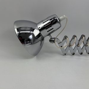 May include: A chrome-finished desk lamp with a flexible, accordion-style arm. The lamp head is a rounded, polished metal, reflecting the surroundings. A white cord extends from the lamp's head. The lamp is designed for focused lighting.
