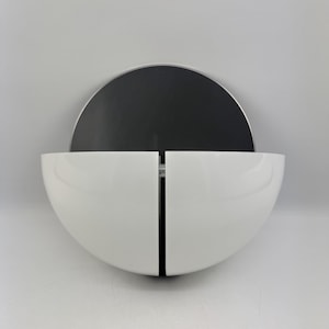 Puede incluir: Una moderna unidad de almacenamiento montada en la pared. El diseño presenta un espejo negro semicircular sobre dos compartimentos blancos en forma de cuenco. Los compartimentos están divididos por una línea negra vertical. La unidad está montada en una pared gris claro.